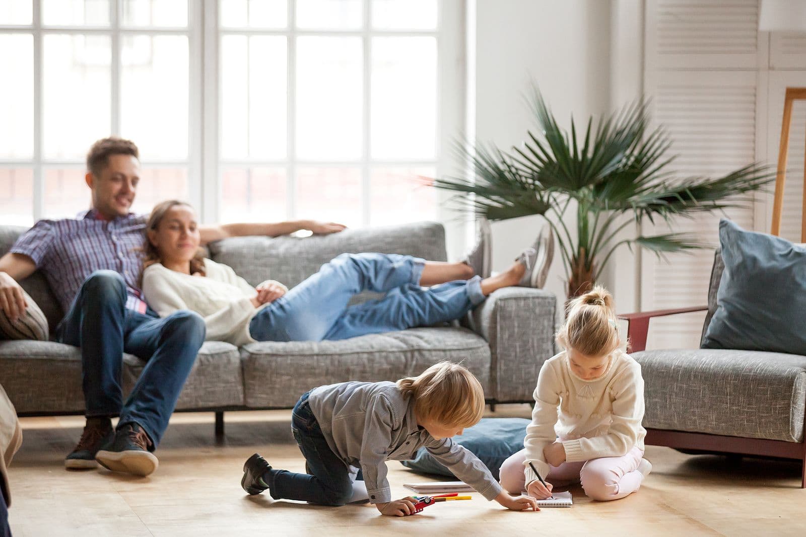 A couple relaxes on a couch while children play on the floor.