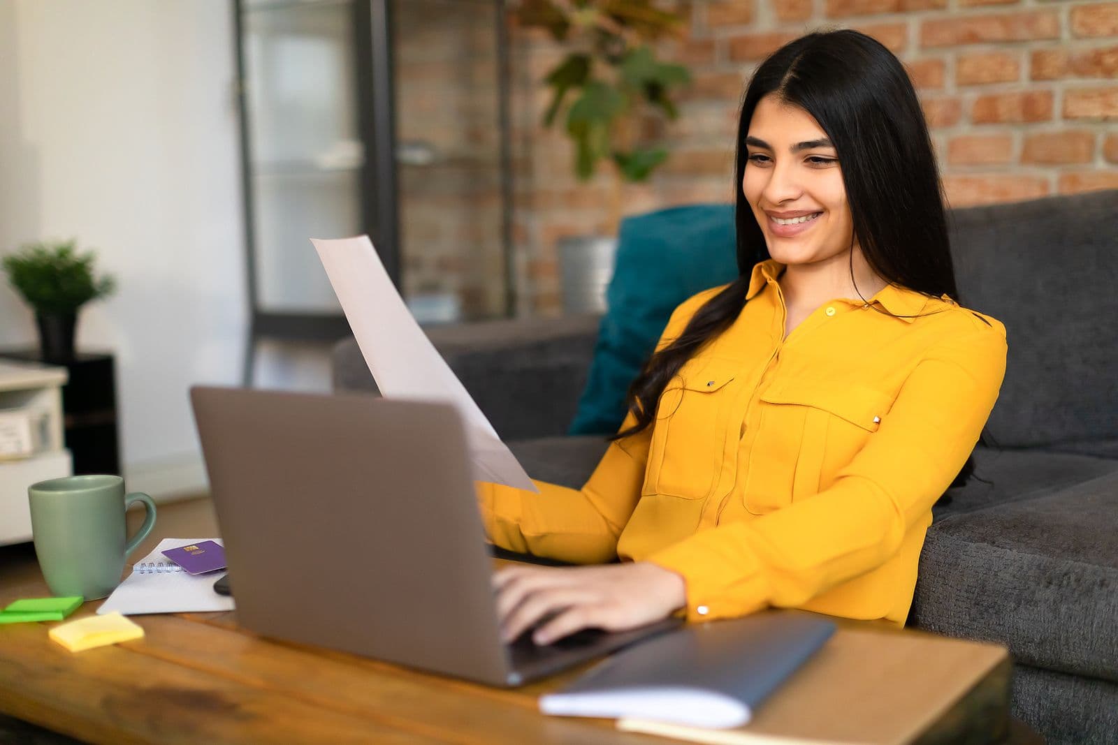 A woman in a yellow shirt sits on the floor between a coffee table and a couch and looks at a laptop.