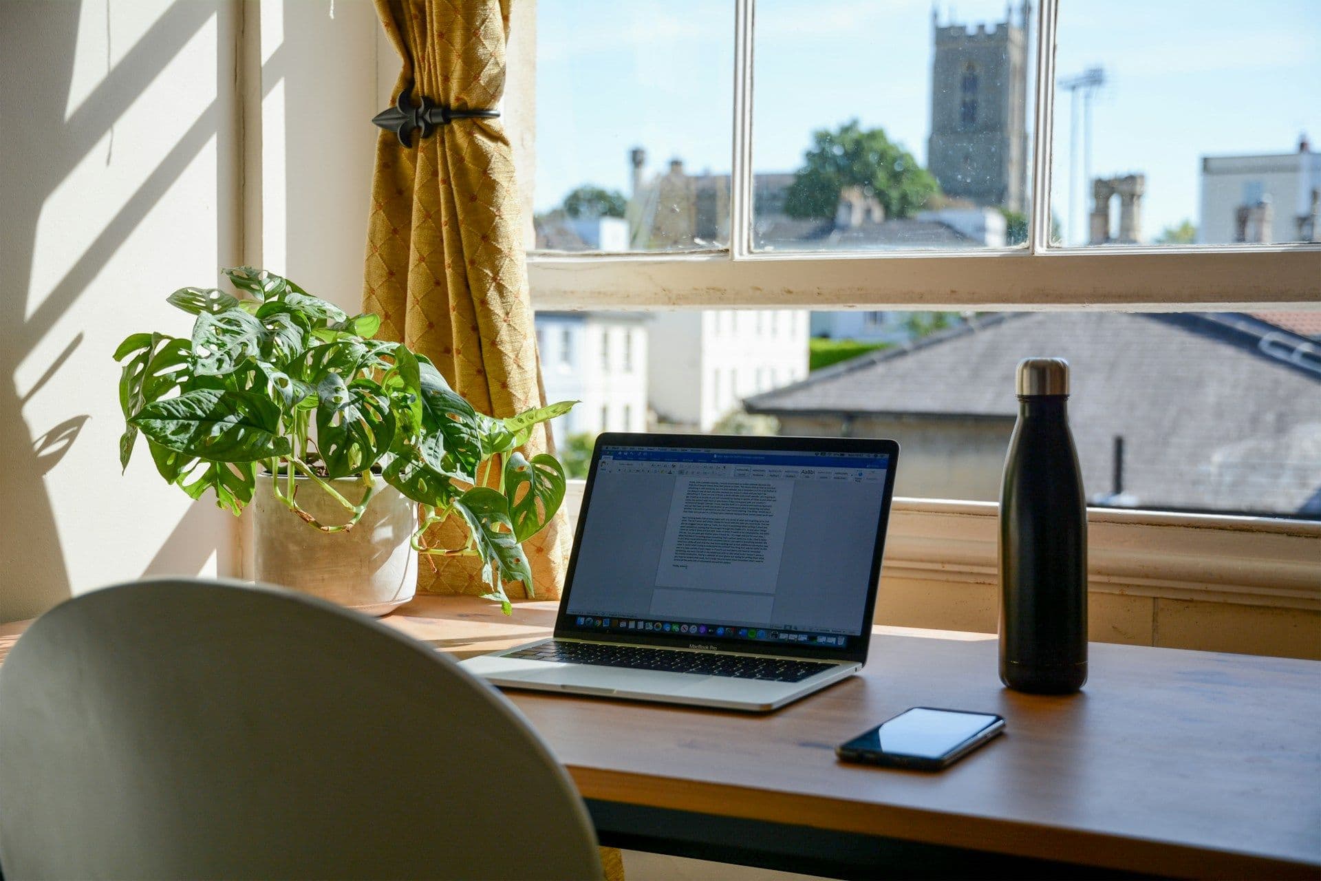 A laptop, phone, and water bottle sit on a desk in front of a window.